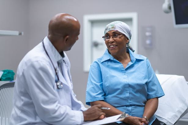 A black senior woman with cancer is at a medical consultation. She is wearing a scarf on her head to hide her hair loss. The doctor is a black man. The patient and doctor are sitting next to each other on an examination table in a medical clinic. The doctor is asking the patient questions and taking notes on a clipboard. The patient is smiling with hope and gratitude.